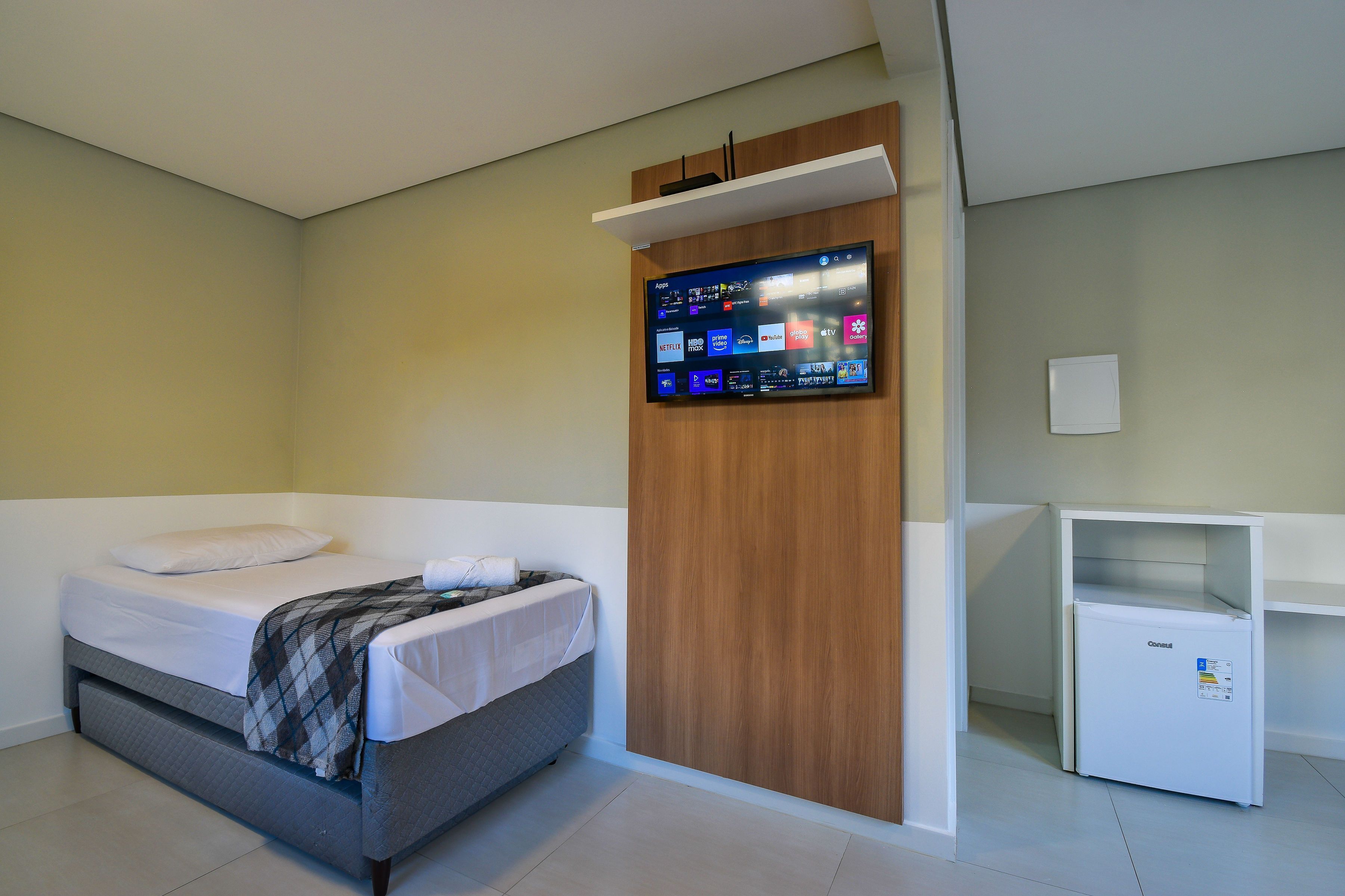 A hotel room with a bed, a flat-screen TV mounted on the wall, and a small white cabinet.