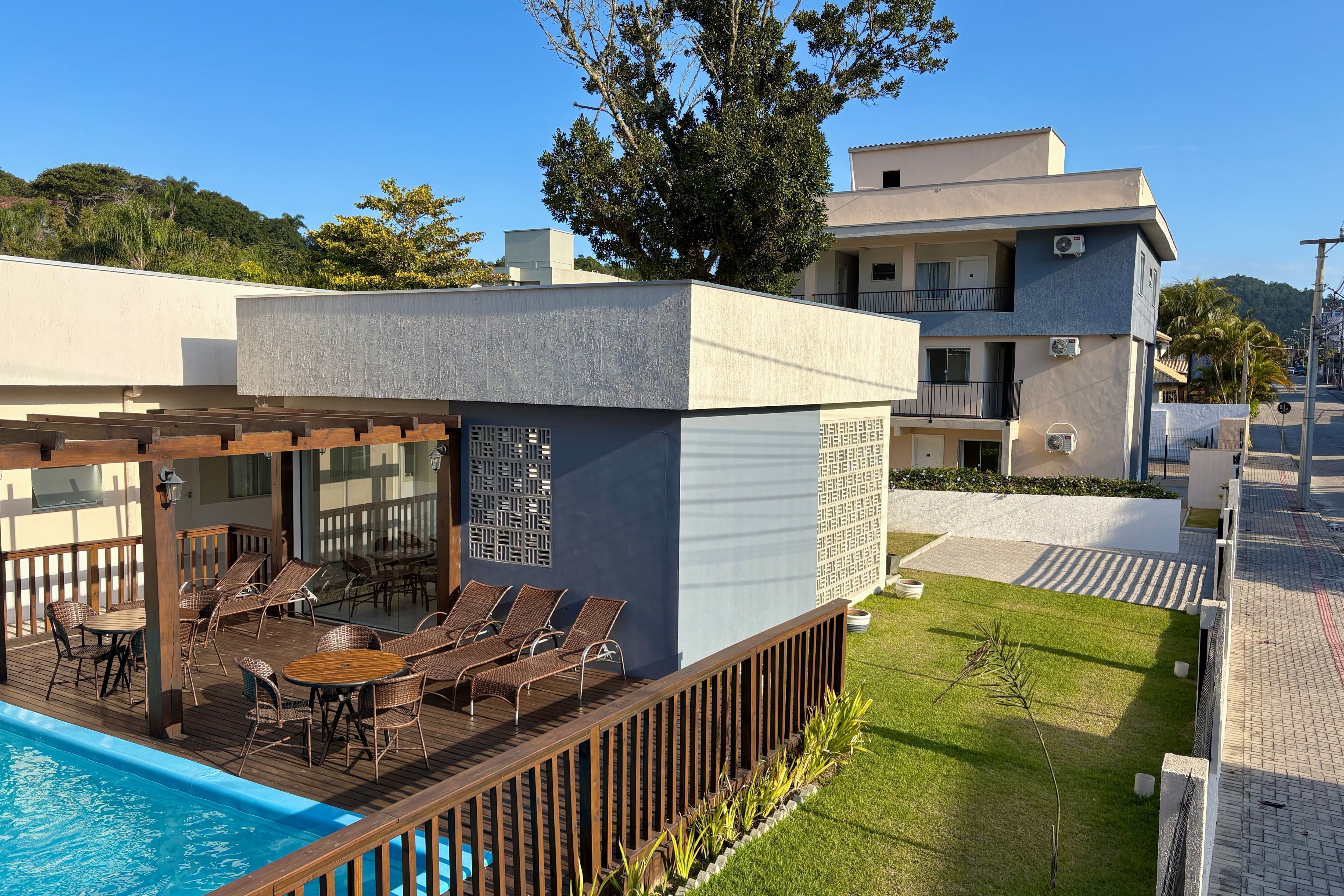 Un espace aménagé au bord de la piscine, avec des chaises longues et une table, entouré d’une terrasse en bois et d’un petit jardin.