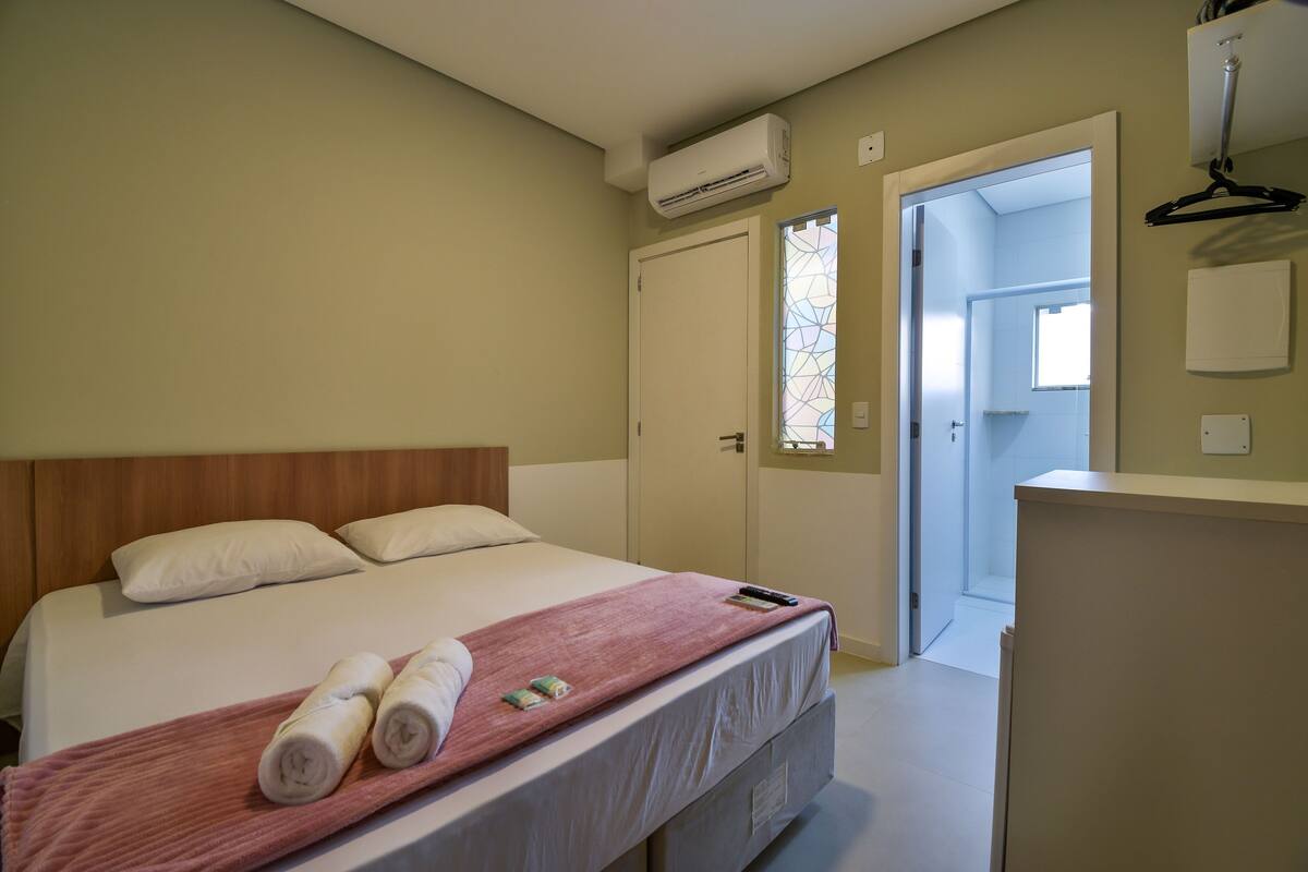 A hotel room with a bed, a pink towel, and a white blanket. There is a door leading to another room and a window with a geometric pattern.