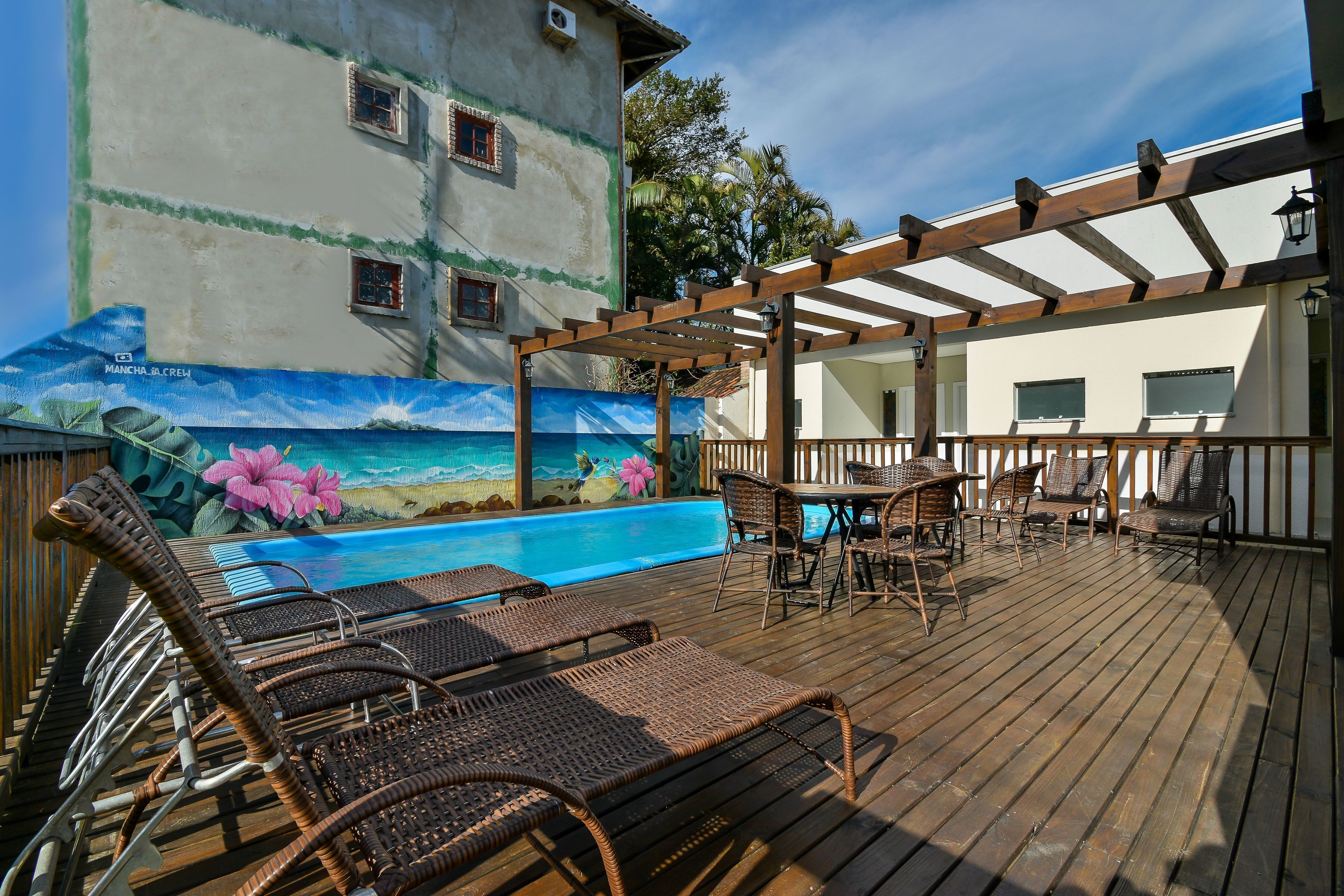Eine Holzterrasse mit Korlounge-Stühlen, ein Pool mit Wandgemälde und eine Pergola.