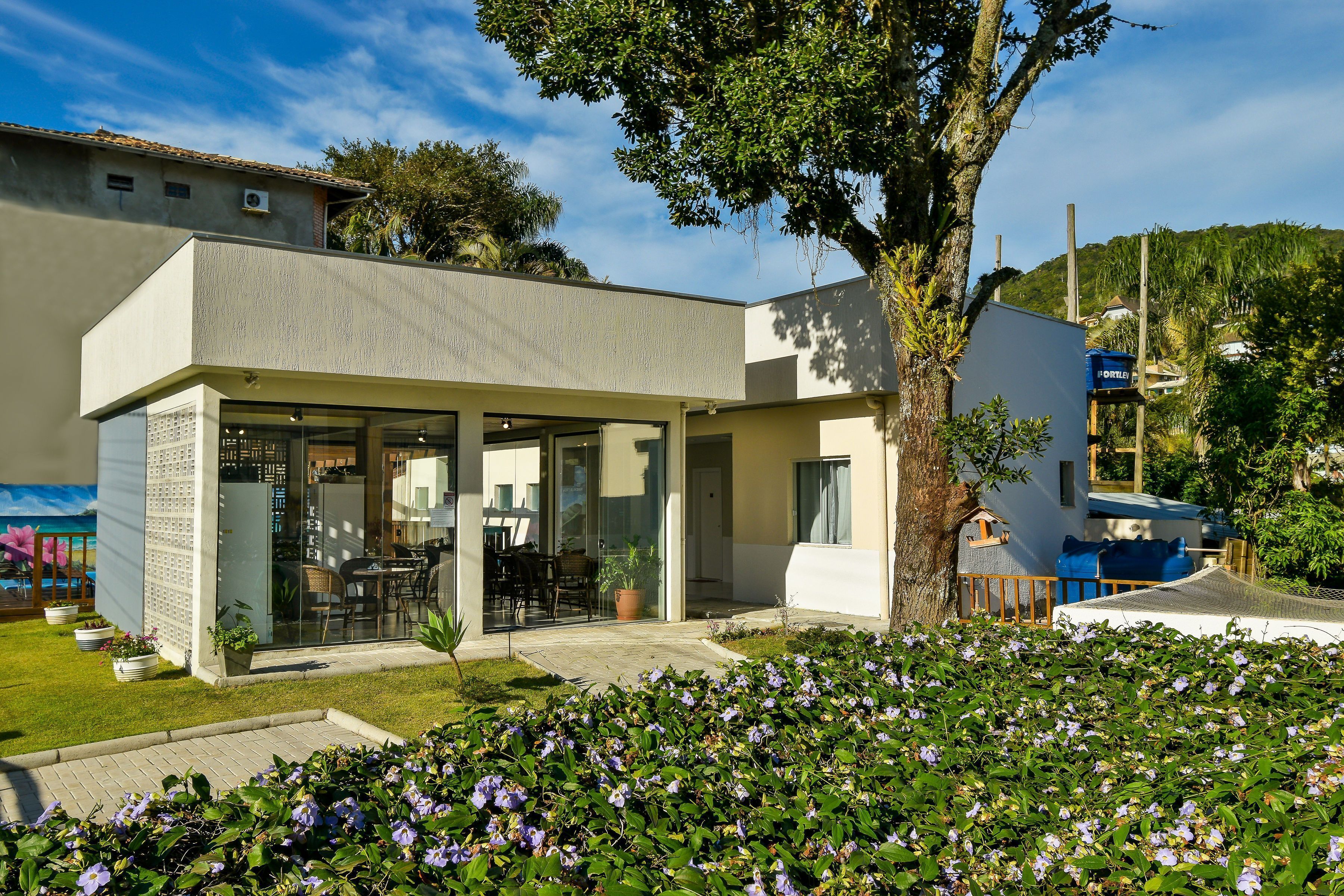 A modern building with a glass entrance, a patio with outdoor seating, and a garden with purple flowers.
