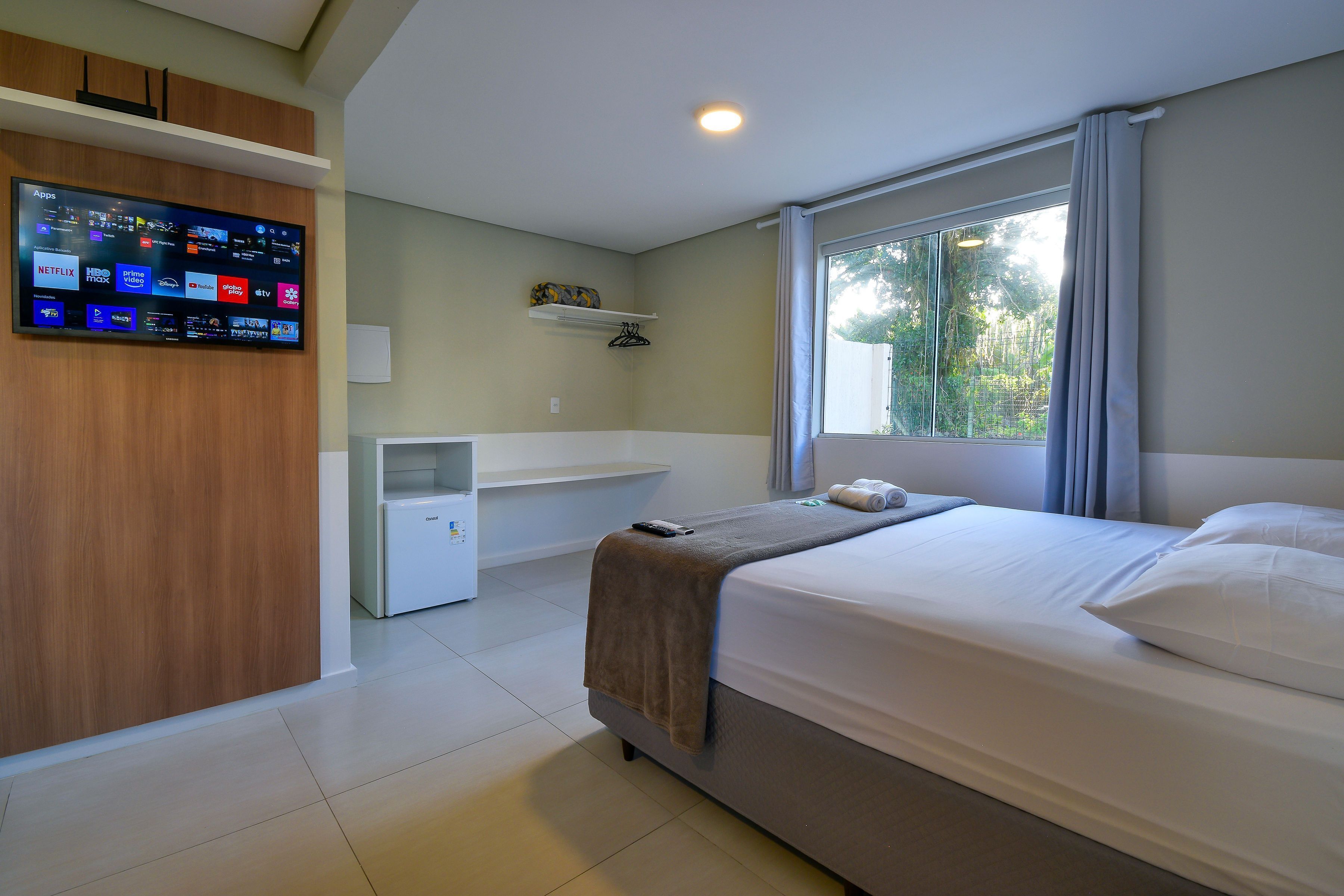 Habitación de hotel con cama, televisor montado en la pared mostrando varias aplicaciones, escritorio y ventana con cortinas.