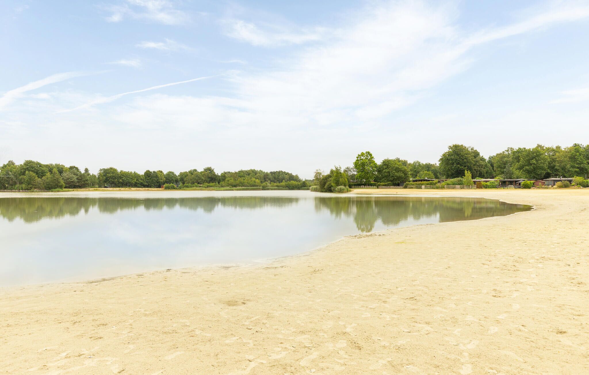Een rustig strand met een kalm meer, omgeven door bomen en enkele gebouwen in de verte.