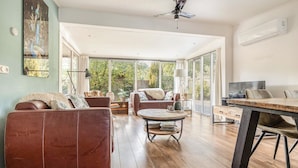 A spacious living room with a wooden floor, leather sofas, a round coffee table, and a dining table with chairs. Large windows provide natural light and views of greenery outside.