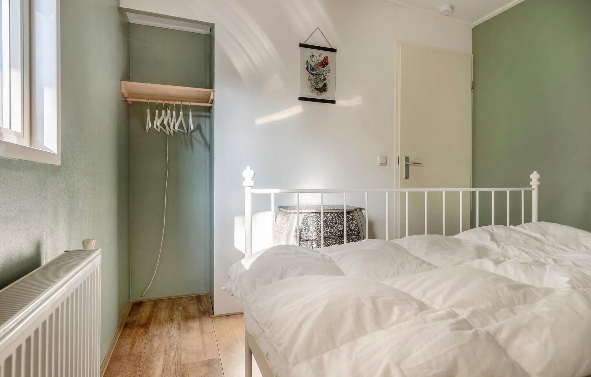 A bedroom with a white bed, a radiator, a wooden shelf, and a hanging laundry rack.