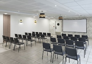 Une salle de conférence avec des rangées de chaises, un écran de projection et un tableau blanc.