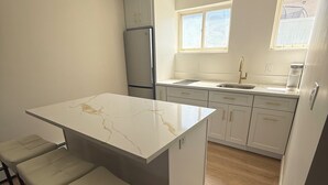 Et moderne køkken med en marmor bordplade, hvide skabe og et rustfrit stålkøleskab. Tre barstole og et vindue giver naturligt lys.