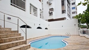 Piscina com deck de pedra, escadas que levam a uma varanda, e um edifício branco com janelas protegidas por grades.