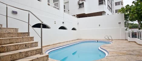 Una piscina con cubierta de piedra, una escalera que lleva a un balcón y un edificio blanco con ventanas protegidas por barrotes.