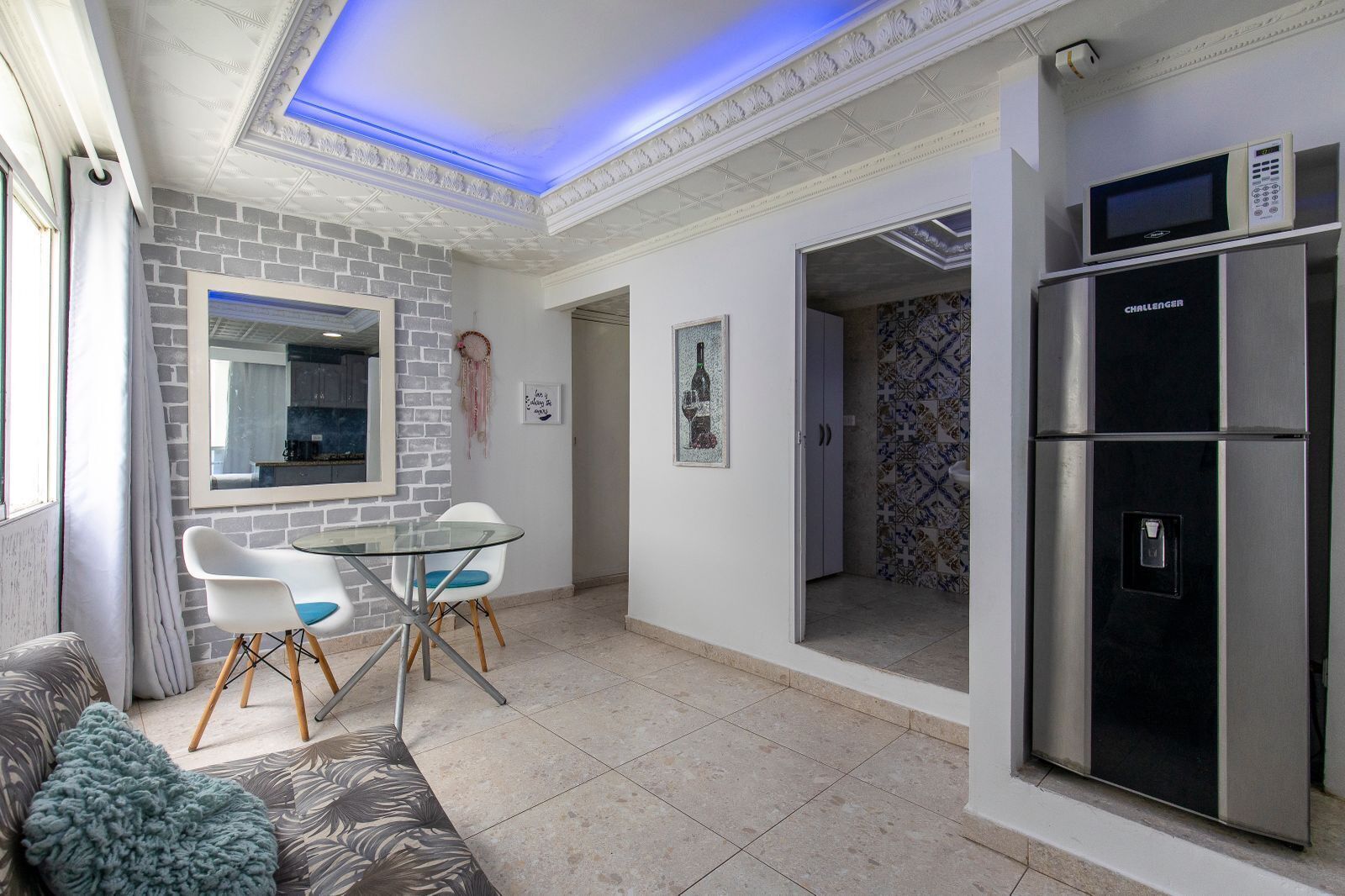 Una sala de estar moderna con una mesa de vidrio, sillas blancas, un sofá y una zona de cocina con refrigerador y microondas.