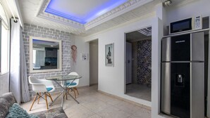 Uma sala moderna com mesa de vidro, cadeiras brancas, um sofá e uma área de cozinha com frigorífico e micro-ondas.