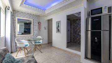 Una sala de estar moderna con una mesa de vidrio, sillas blancas, un sofá y una zona de cocina con refrigerador y microondas.