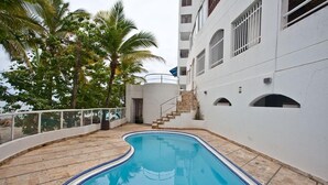 Una piscina con solárium de piedra, rodeada de palmeras y un edificio blanco con ventanas de arco y una escalera que conduce a un nivel superior.