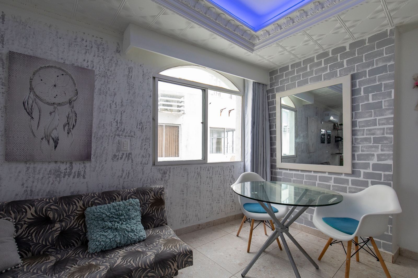 Una sala de estar con un sofá estampado, una mesa de vidrio con sillas, un espejo grande y un atrapasueños en la pared.
