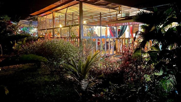 A well-lit, modern building with a glass facade and a covered outdoor area, surrounded by lush greenery. - Las Casitas italiana (Sabalito)