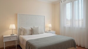 A neatly arranged bedroom with a large bed, two bedside tables with lamps, a headboard with a leaf pattern, and sheer curtains on the window.