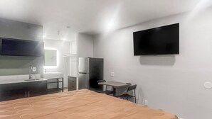 A compact hotel room with a bed, a small desk, a refrigerator, a flat-screen TV mounted on the wall, and a window providing natural light. - Value Suites Extended Stay (Sacramento)