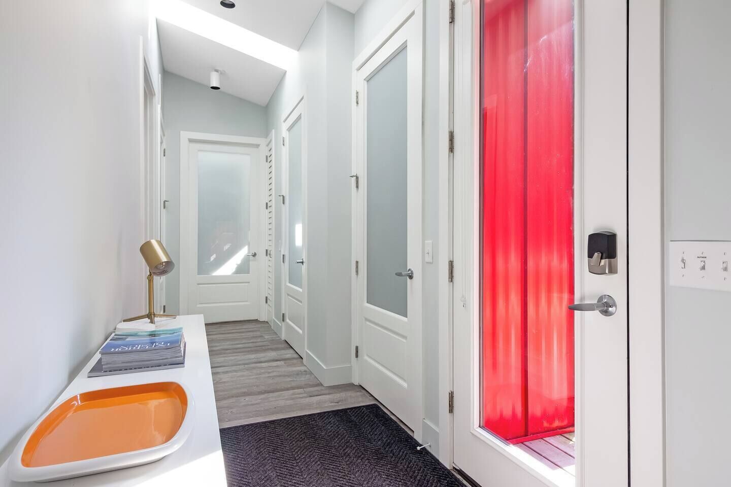 A hallway with a white door, a red door, and a small table with a lamp and magazines.