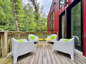 A deck with white chairs and yellow tables, surrounded by a wooden deck and a red building with large windows.