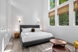 A bedroom with a large bed, bedside lamps, a round table, and a view of trees through the windows.