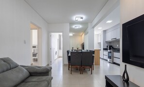 Ein modernes Wohnzimmer mit grauem Sofa, einem Essbereich mit Tisch und Stühlen, einer Küche mit Geräten und einem an der Wand montierten Fernseher.