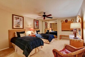 A hotel room with two beds, a ceiling fan, a mirror, a bedside table with a lamp, and a wicker chair.