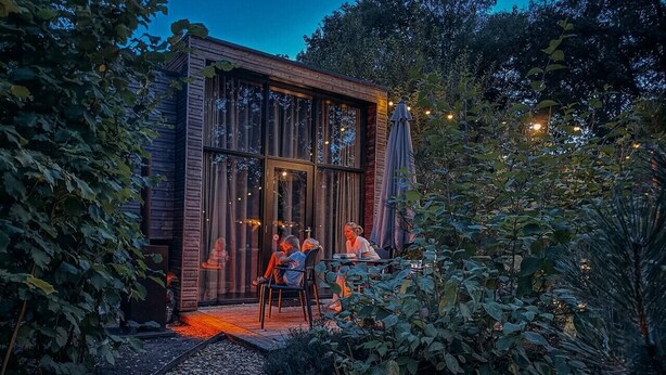 A wooden cabin with large windows, a patio with chairs, and a garden path.