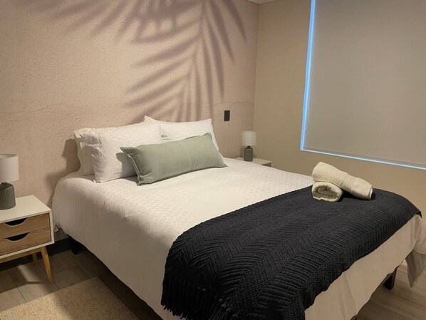 Una cama bien hecha con almohadas blancas y verdes, un plaid oscuro y una toalla doblada al lado.