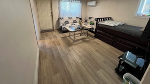 A compact room with a bed, a desk, and a chair. There is a window with curtains, a door, and an air conditioning unit on the wall.