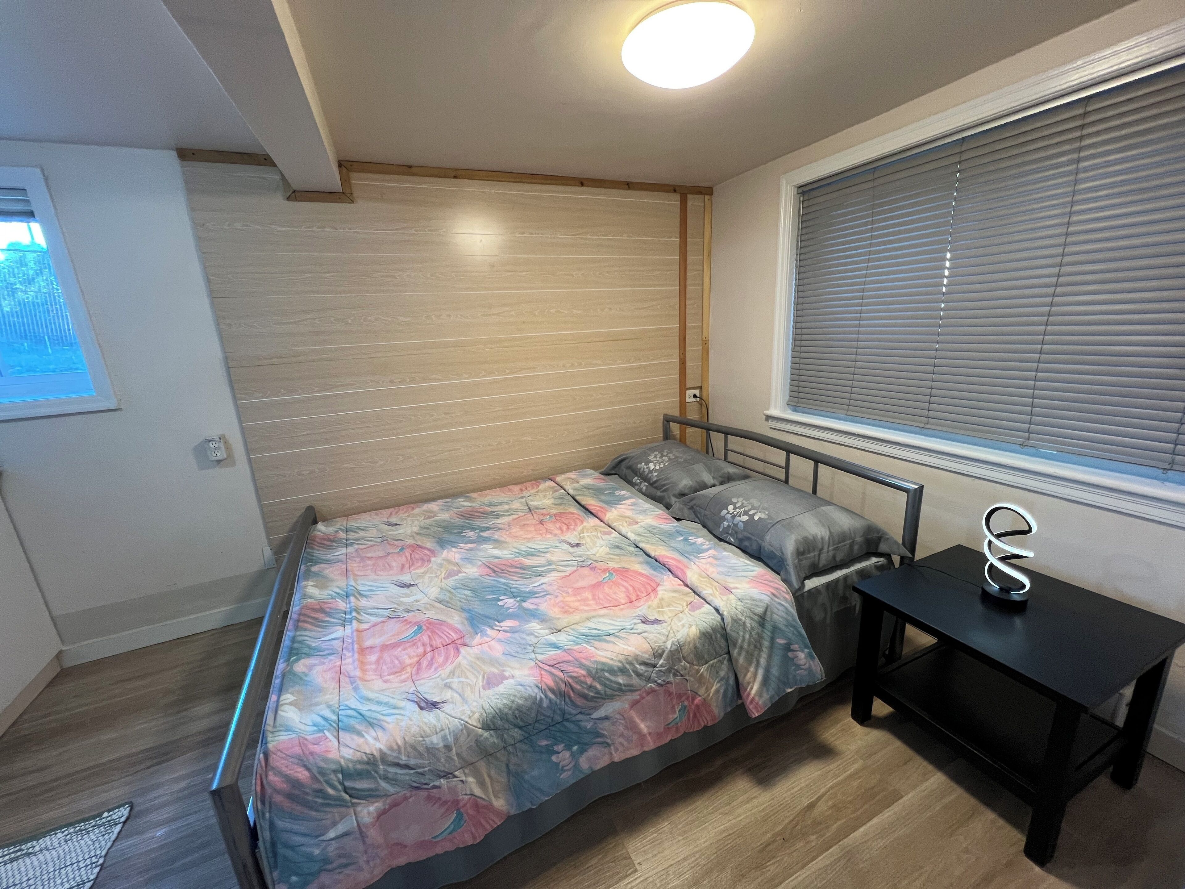 A bedroom with a bed, a small table, a window with blinds, and a ceiling light.