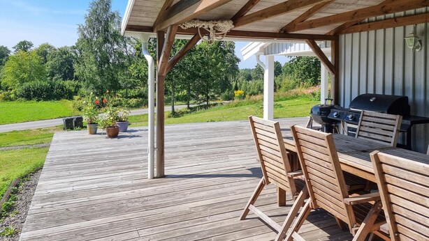 An outdoor patio with wooden furniture and a grill, surrounded by greenery and a clear view of the landscape. - Awesome home in Värnamo with lake view (Värnamo)