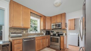 A modern kitchen with wooden cabinets, stainless steel appliances, and a large window providing natural light. - 5485-Hammerhead Harbour, Old Nags Head Cove | Pet-Friendly Coastal Escape (Nags Head)