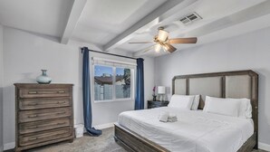 Ein Schlafzimmer mit Bett, Schminktisch, Nachttisch, Deckenventilator und einem Fenster mit Vorhängen.