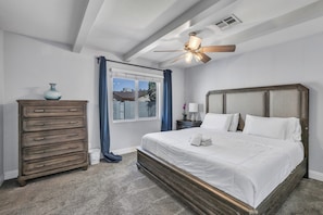 Une chambre à coucher comprenant un lit, une commode, une table de chevet, un ventilateur de plafond et une fenêtre avec des rideaux.
