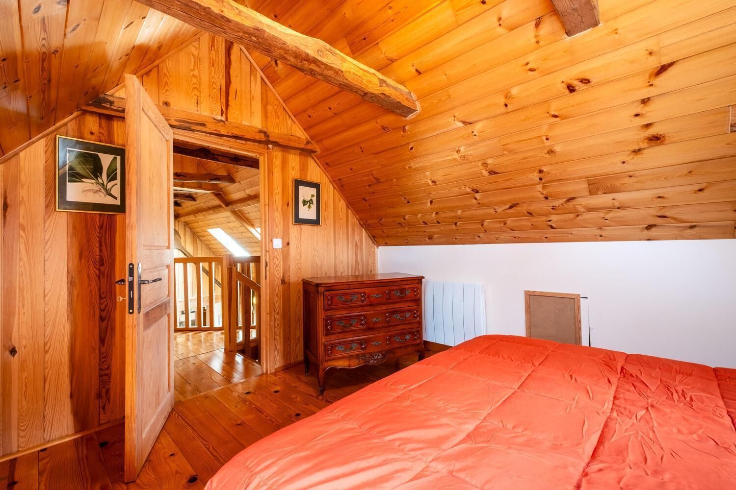 A wooden room with a sloped ceiling, a bed with a red blanket, a dresser, and two framed pictures on the wall.