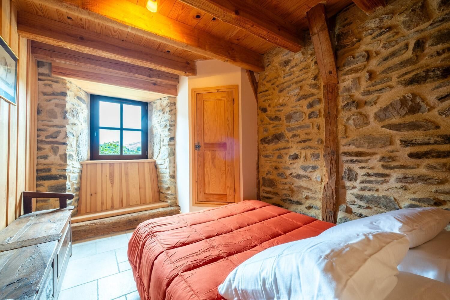 A bedroom with a stone wall, wooden beams, a wooden bench, and a window.