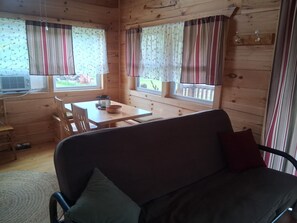 A wooden cabin interior with a sofa, a dining table, and two windows with curtains.
