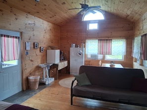 A wooden cabin interior with a kitchenette, a sofa, a ceiling fan, and a window with curtains.