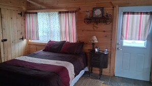 A wooden cabin bedroom with a bed, curtains, a small table with a lamp, and a door. - Private cabin on the beautiful Kickapoo River Valley near Wildcat Mountain Park. (Ontario)
