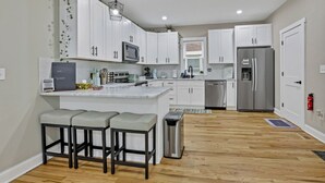A modern kitchen with white cabinets, a stainless steel refrigerator, and a bar area with stools. - The Vibe at OSU: Walking distance to the stadium! (Columbus)