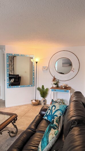 A living room with a brown leather sofa, a large mirror with a seashell frame, a floor lamp, and a small table with decorative items.