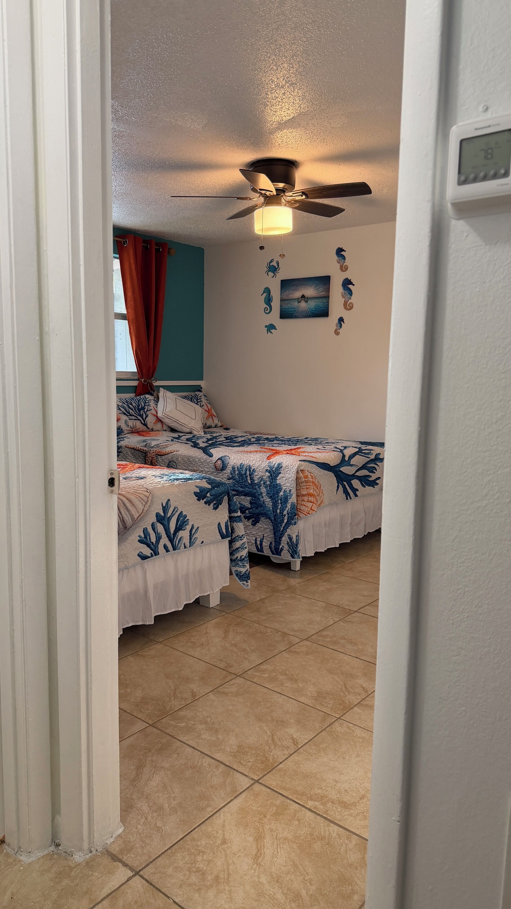 Une chambre avec deux lits, un ventilateur de plafond et un thermostat fixé au mur.