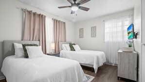 A hotel room with two beds, a ceiling fan, a TV, and a small desk with a lamp.