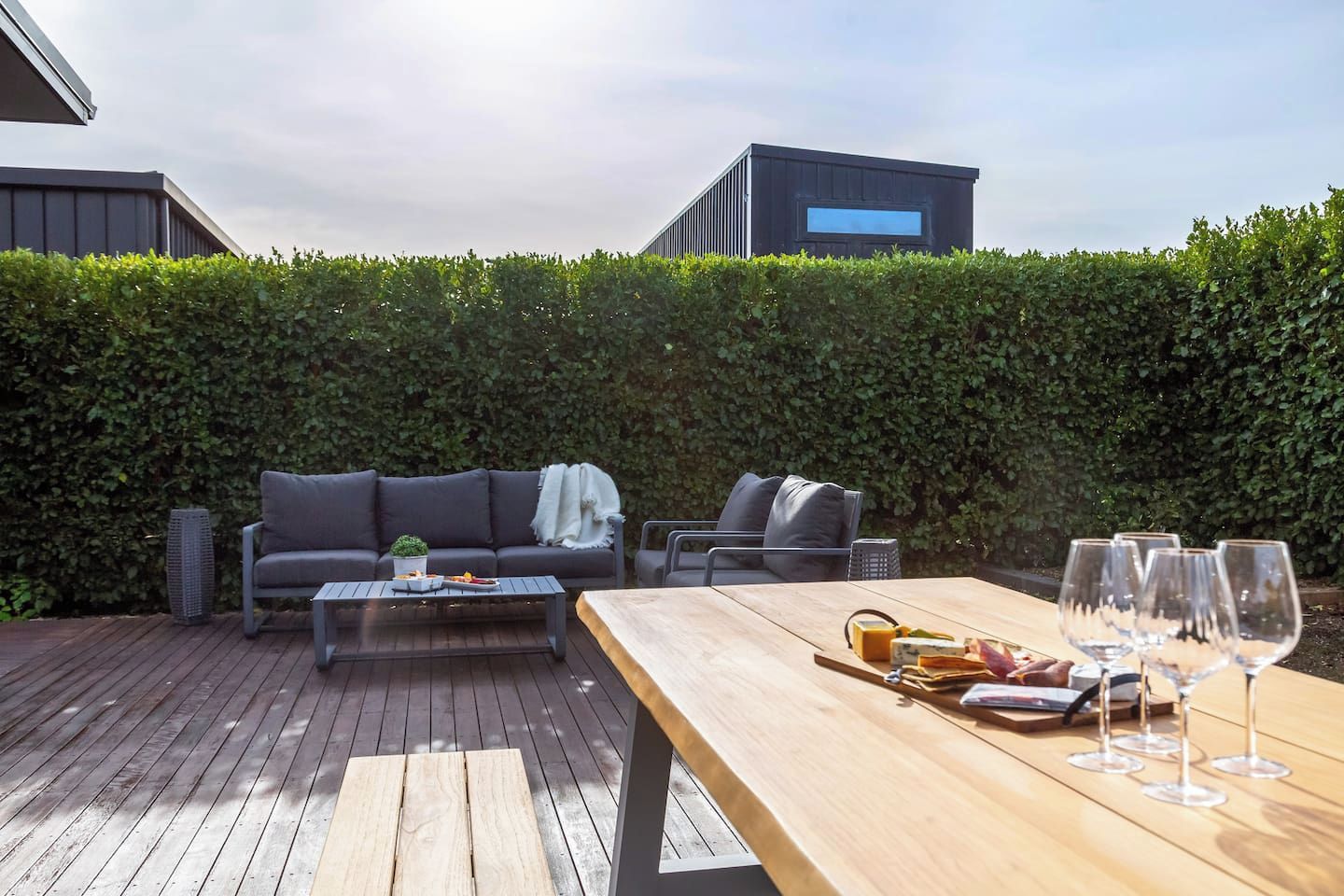 Un patio exterior con terraza de madera, un conjunto de sofás, una mesa de comedor con copas de vino y un seto al fondo.