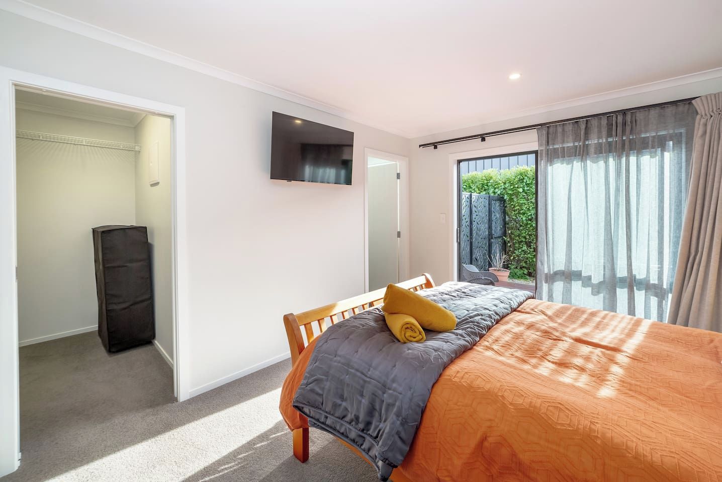 A bedroom with a bed, a TV mounted on the wall, a closet, and a view of greenery outside.