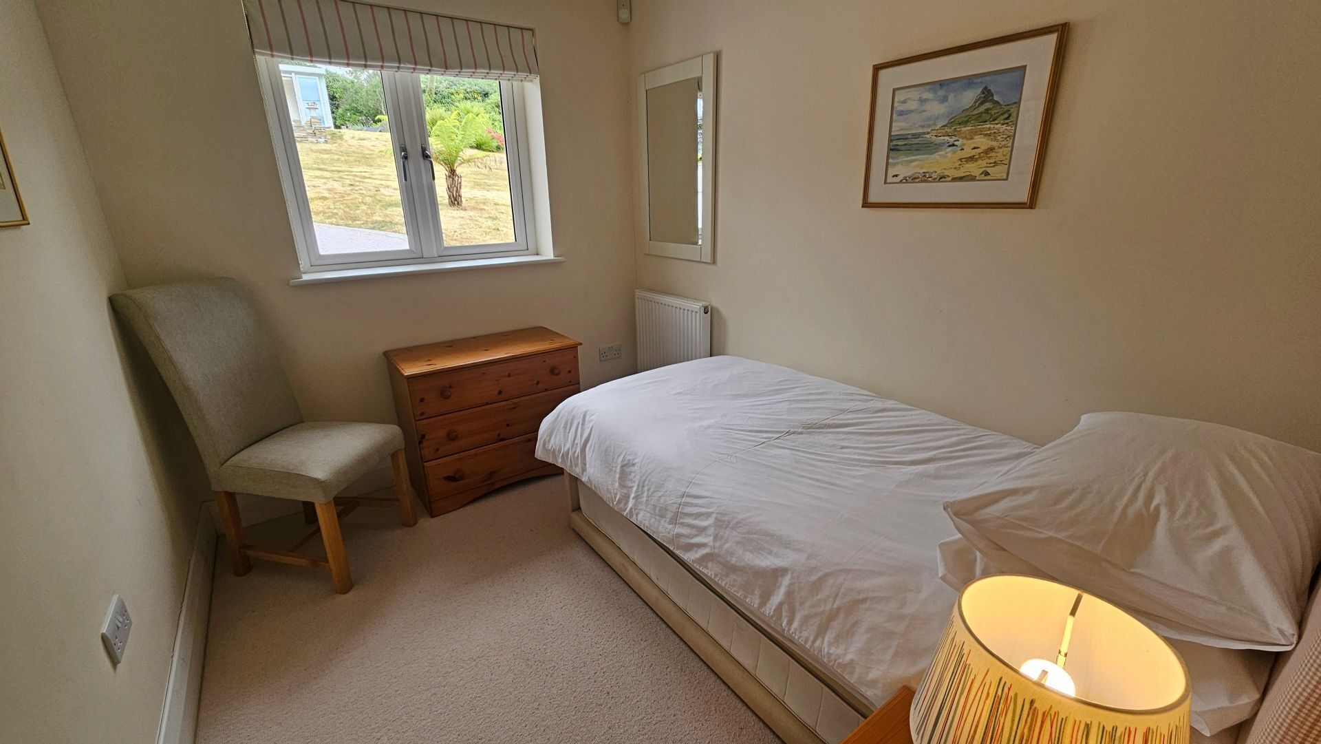 A bedroom with a single bed, a chair, a dresser, a window with a view of greenery, and a painting on the wall.