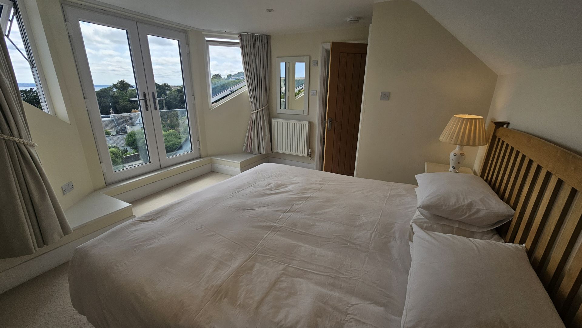 A bedroom with a large bed, a window with curtains, a wooden headboard, and a nightstand with a lamp.