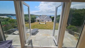 A view through a glass door of a house with a stone patio, a lawn, and a distant body of water. - Highlands - sleeps 7 guests  in 4 bedrooms (St Mawes)