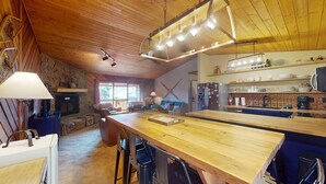 Interior acogedor de una cabaña con mesa de comedor de madera, taburetes de bar, chimenea y área de cocina con estantes y electrodomésticos.