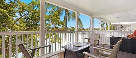 A covered outdoor area with a wooden deck, a table with chairs, and a view of palm trees and a house in the background.